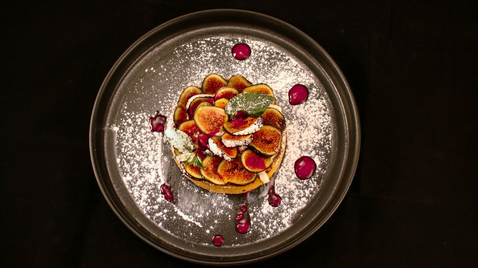 Delicious fig tart displayed with powdered sugar and sauce on a dark plate.